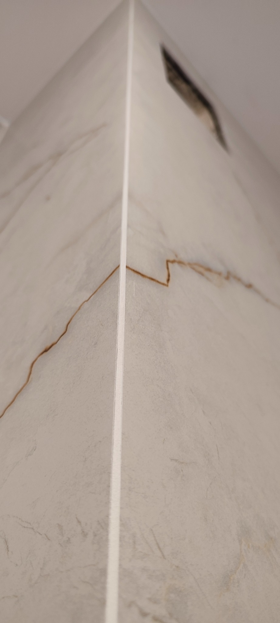 Detal narożnika wykończonego płytkami imitującymi marmur z fugą. Widoczny fragment kratki wentylacyjnej w górnej części.