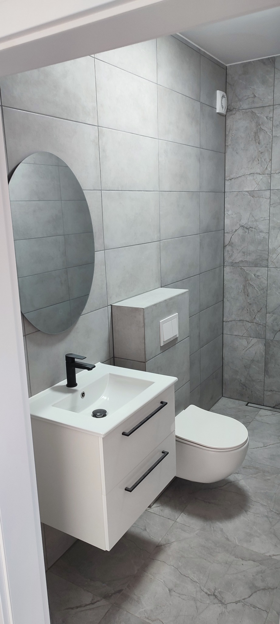 Nowoczesna łazienka z szarymi płytkami, okrągłym lustrem i białą szafką pod umywalką z czarną armaturą. Minimalistyczny design z wiszącą toaletą i subtelnym oświetleniem.