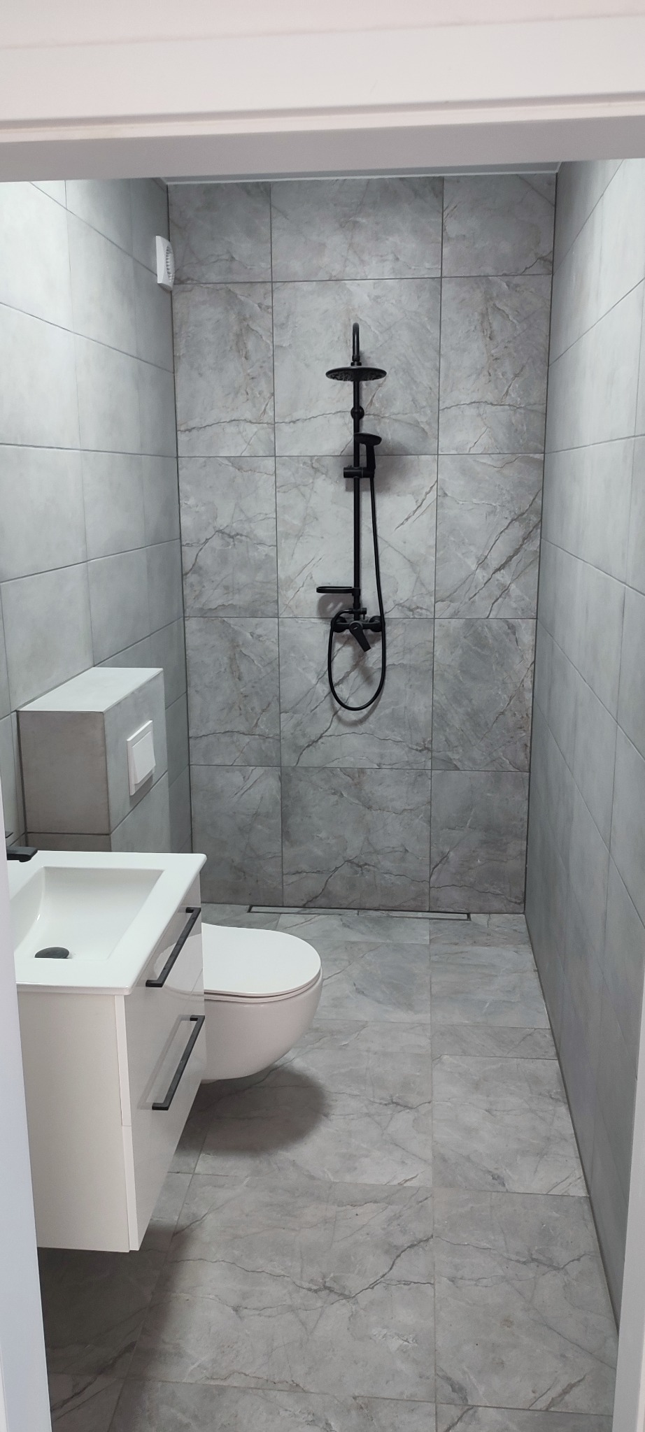 Nowoczesna łazienka z szarymi płytkami imitującymi marmur, białą szafką z umywalką i czarną armaturą prysznicową. Minimalistyczny design i stonowana kolorystyka.