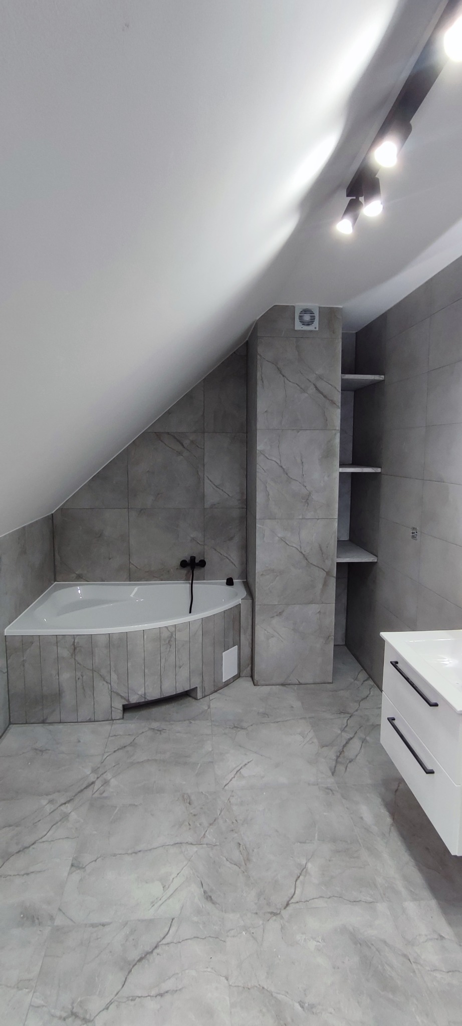 Jasna łazienka na poddaszu z wanną narożną obudowaną płytkami imitującymi marmur, półkami w ścianie i szafką pod umywalką. Oświetlenie sufitowe.