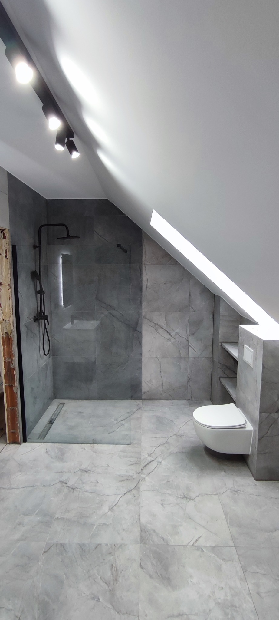 Wykończona łazienka na poddaszu z szarymi płytkami imitującymi marmur, kabiną prysznicową typu walk-in i podwieszaną toaletą. Widoczne oświetlenie punktowe.