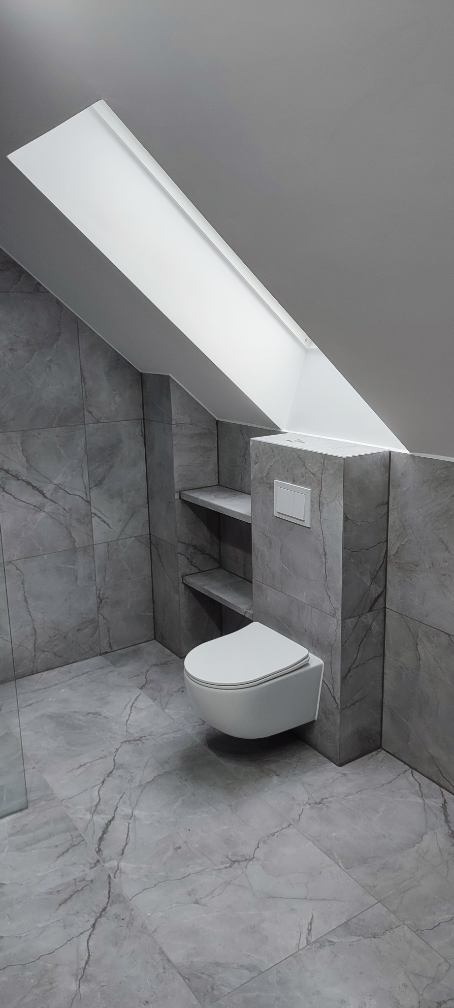 Nowoczesna łazienka na poddaszu z szarą marmurową glazurą, podwieszaną toaletą i półkami w ścianie. Okno dachowe doświetla wnętrze.