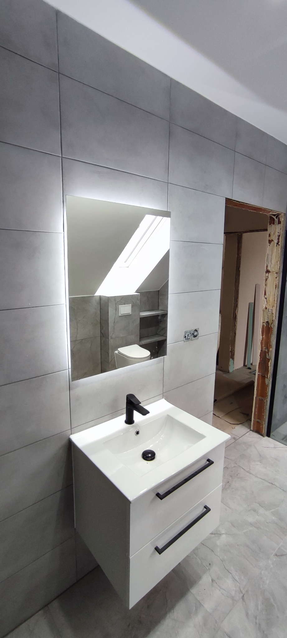 Wykończona łazienka w szarości. Umywalka z szafką i czarną armaturą. W lustrze odbicie toalety i okna dachowego.  Widoczny fragment otwartych drzwi do innego pomieszczenia.