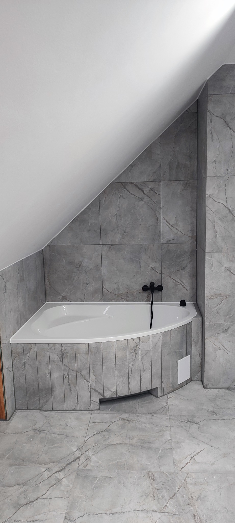 Wykończona łazienka na poddaszu ze skośnym sufitem. Szare płytki imitujące marmur na ścianach i podłodze, biała narożna wanna z czarną armaturą.