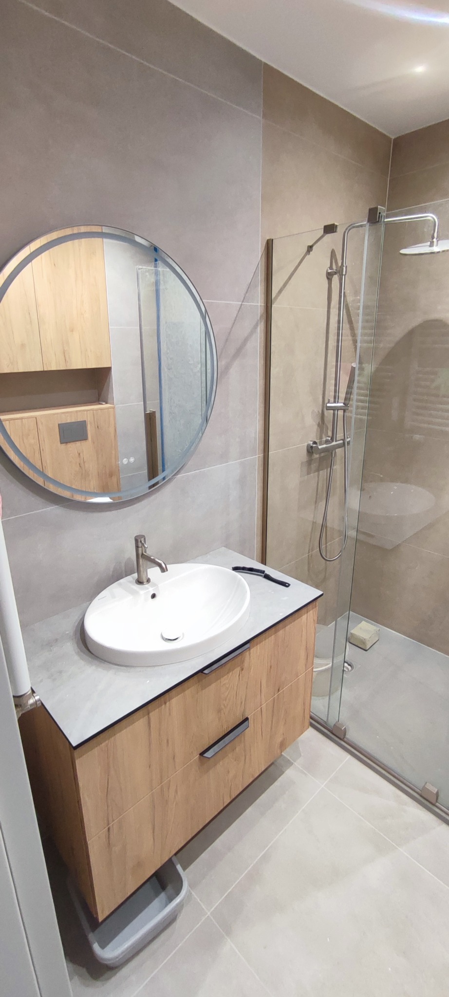Nowoczesna łazienka z kabiną prysznicową, umywalką na szafce z drewnianym frontem i okrągłym lustrem. Szare płytki na ścianach i podłodze. Minimalistyczny design.