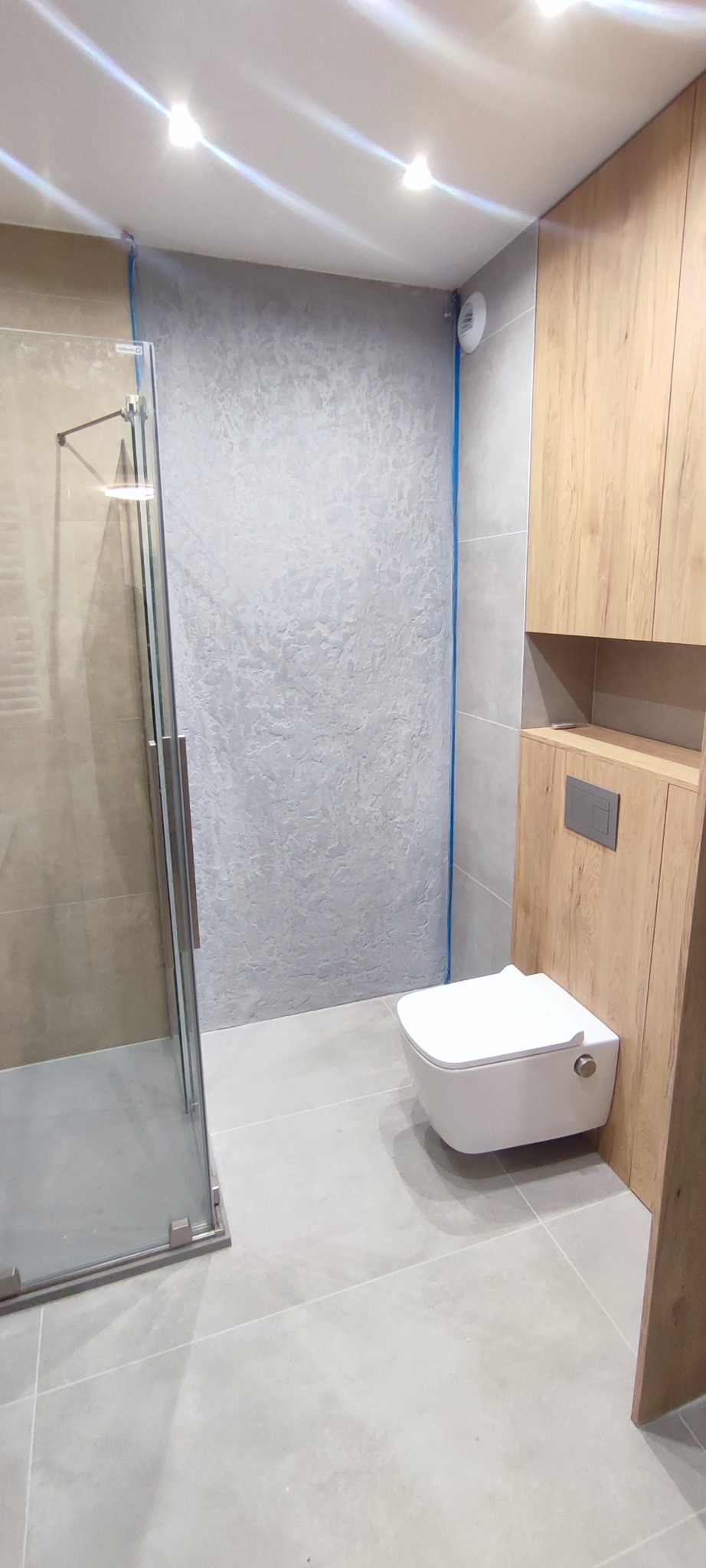Nowoczesna łazienka z kabiną prysznicową, podwieszaną toaletą i drewnianą zabudową. Ściana pokryta dekoracyjną szarą strukturą. Jasne płytki podłogowe.