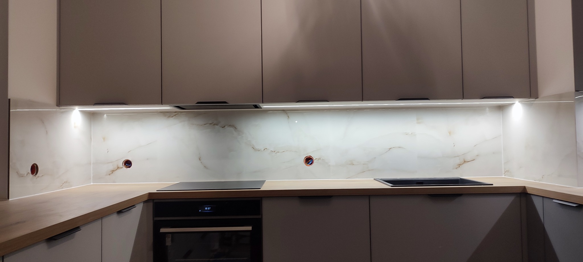 Nowoczesna kuchnia z marmurowym panelem ściennym, podświetleniem LED i minimalistycznymi szafkami w odcieniach szarości. Widoczne gniazdka elektryczne.