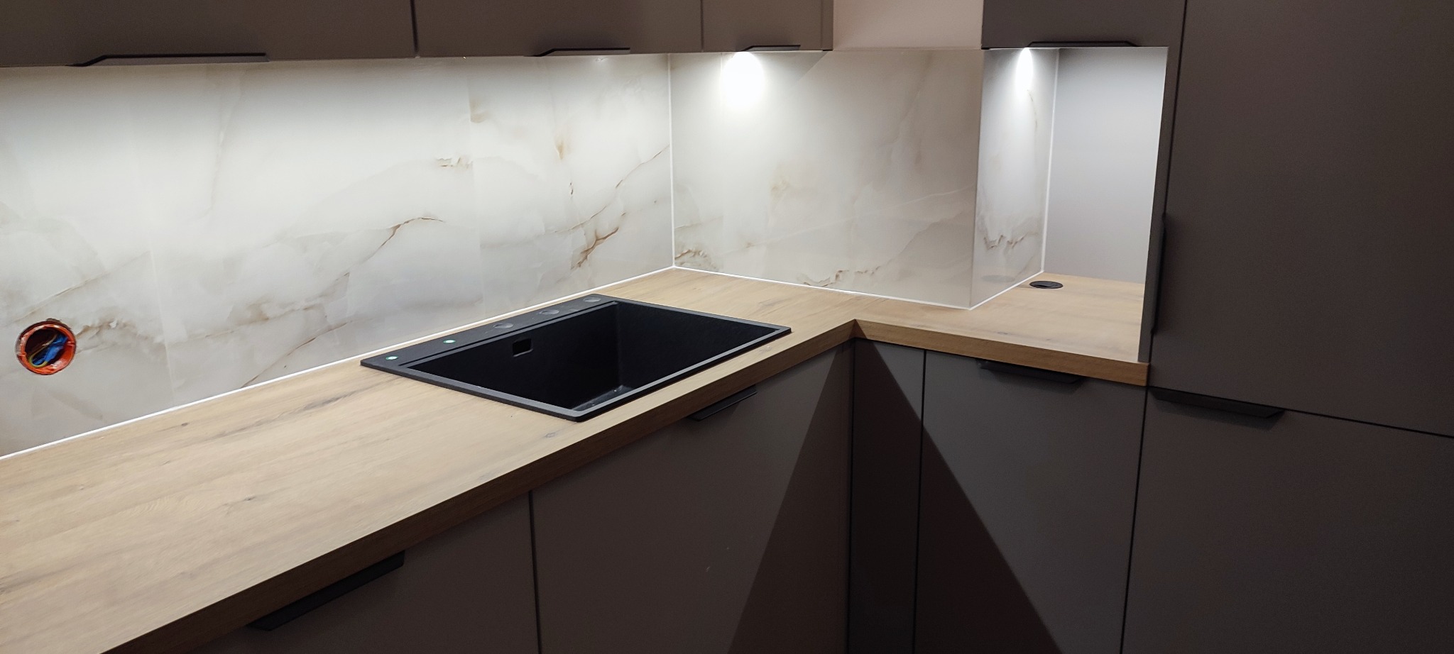 Nowoczesna kuchnia z marmurowymi płytkami nad blatem i czarnym zlewem. Widoczne szare szafki z minimalistycznymi uchwytami i oświetlenie LED pod szafkami.