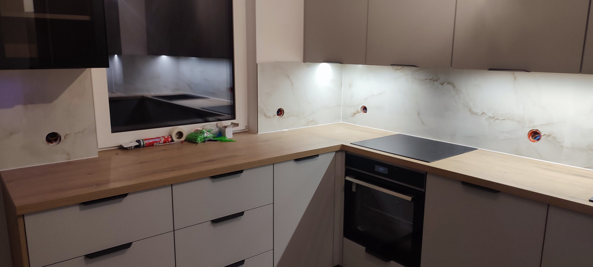 Nowoczesna kuchnia z marmurowym panelem ściennym, drewnianym blatem i szarymi szafkami. Widoczne gniazdka elektryczne i akcesoria montażowe na blacie.