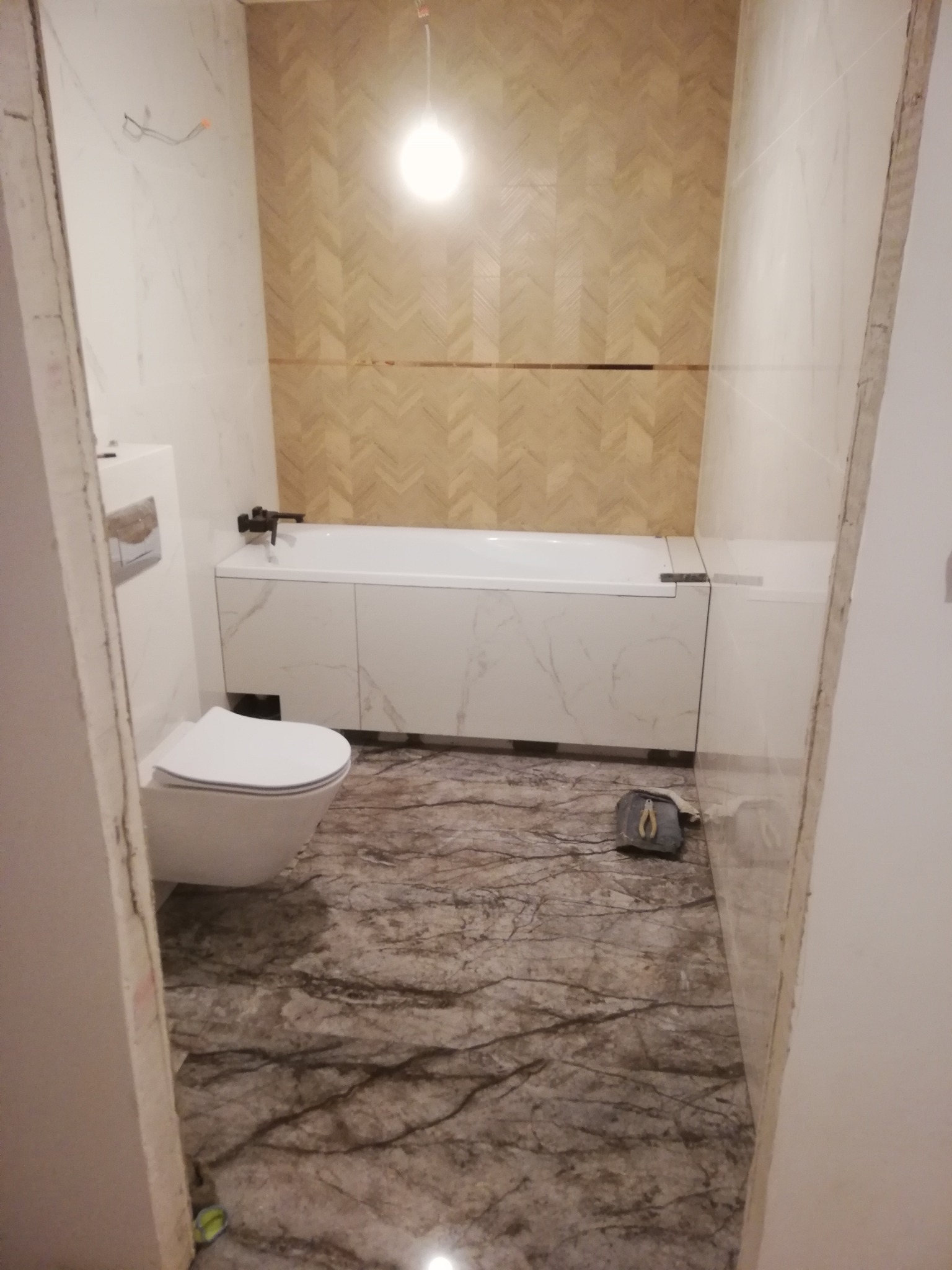 Wykończona łazienka z marmurową podłogą i ścianami. Nad wanną dekoracyjna ściana z wzorem jodełki. Widoczny fragment toalety i narzędzia glazurnicze.