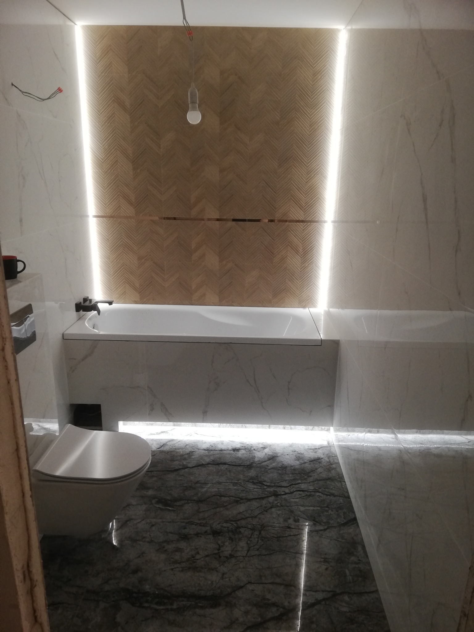 Nowoczesna łazienka z marmurowymi płytkami, drewnianą mozaiką na ścianie i podświetleniem LED. Widoczna wanna, toaleta i fragment instalacji elektrycznej.