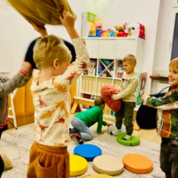 Aktywna zabawa w grupie dzieci w jasnym pomieszczeniu. Dzieci bawią się poduszkami i kolorowymi podestami, rozwijając motorykę i wsp&oacute;łpracę. Radosna atmosfera.