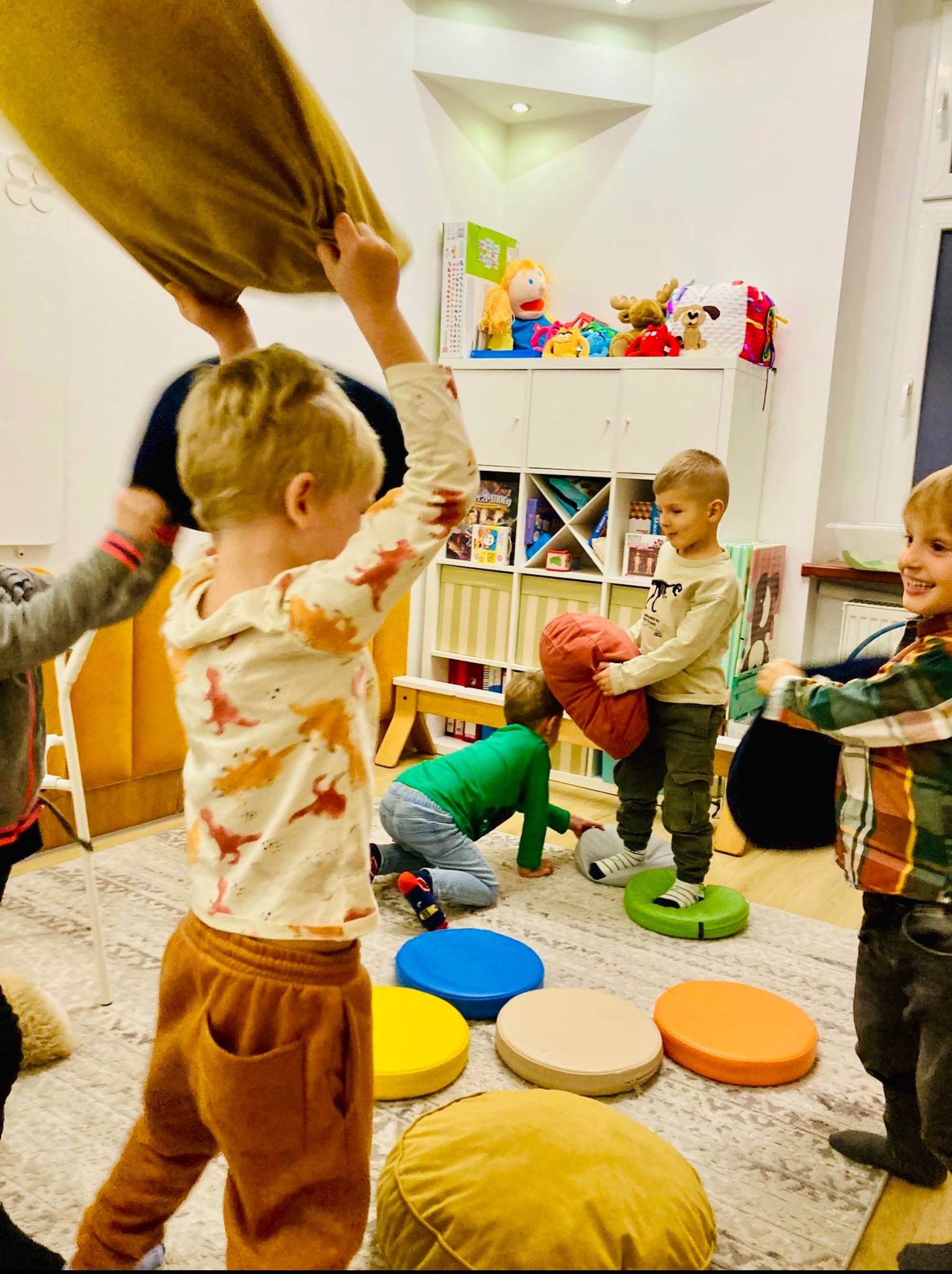 Aktywna zabawa w grupie dzieci w jasnym pomieszczeniu. Dzieci bawią się poduszkami i kolorowymi podestami, rozwijając motorykę i współpracę. Radosna atmosfera.