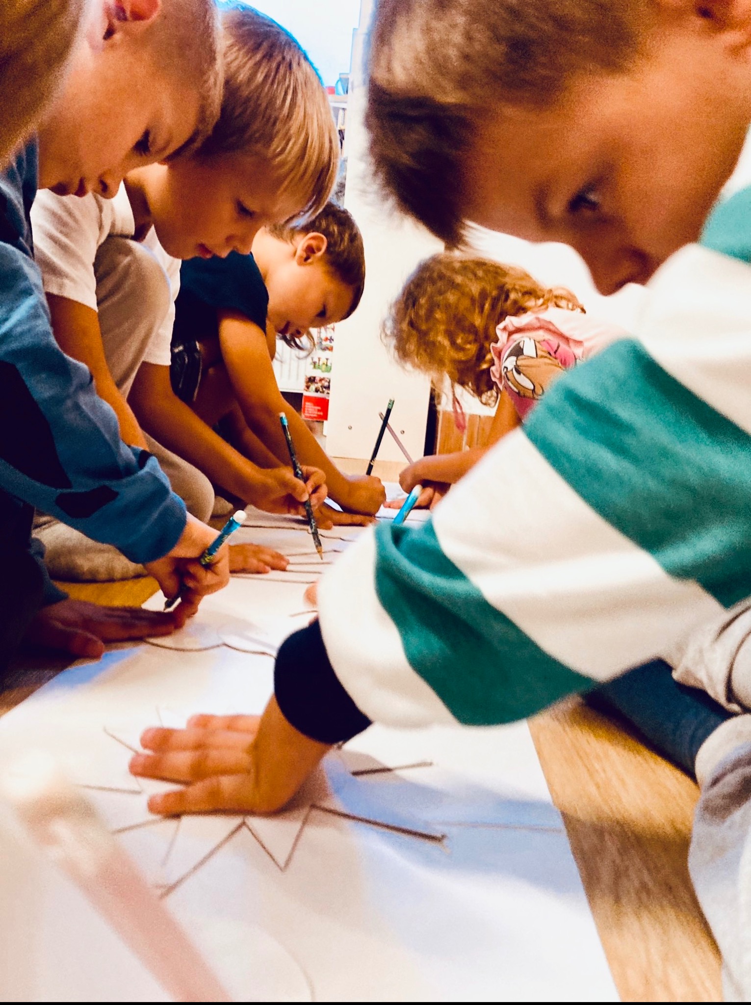 Grupa dzieci pochylona nad dużym arkuszem, rysująca wspólnie gwiazdę. Używają kolorowych kredek, skupione na zadaniu, widać ich dłonie i fragmenty ubrań.