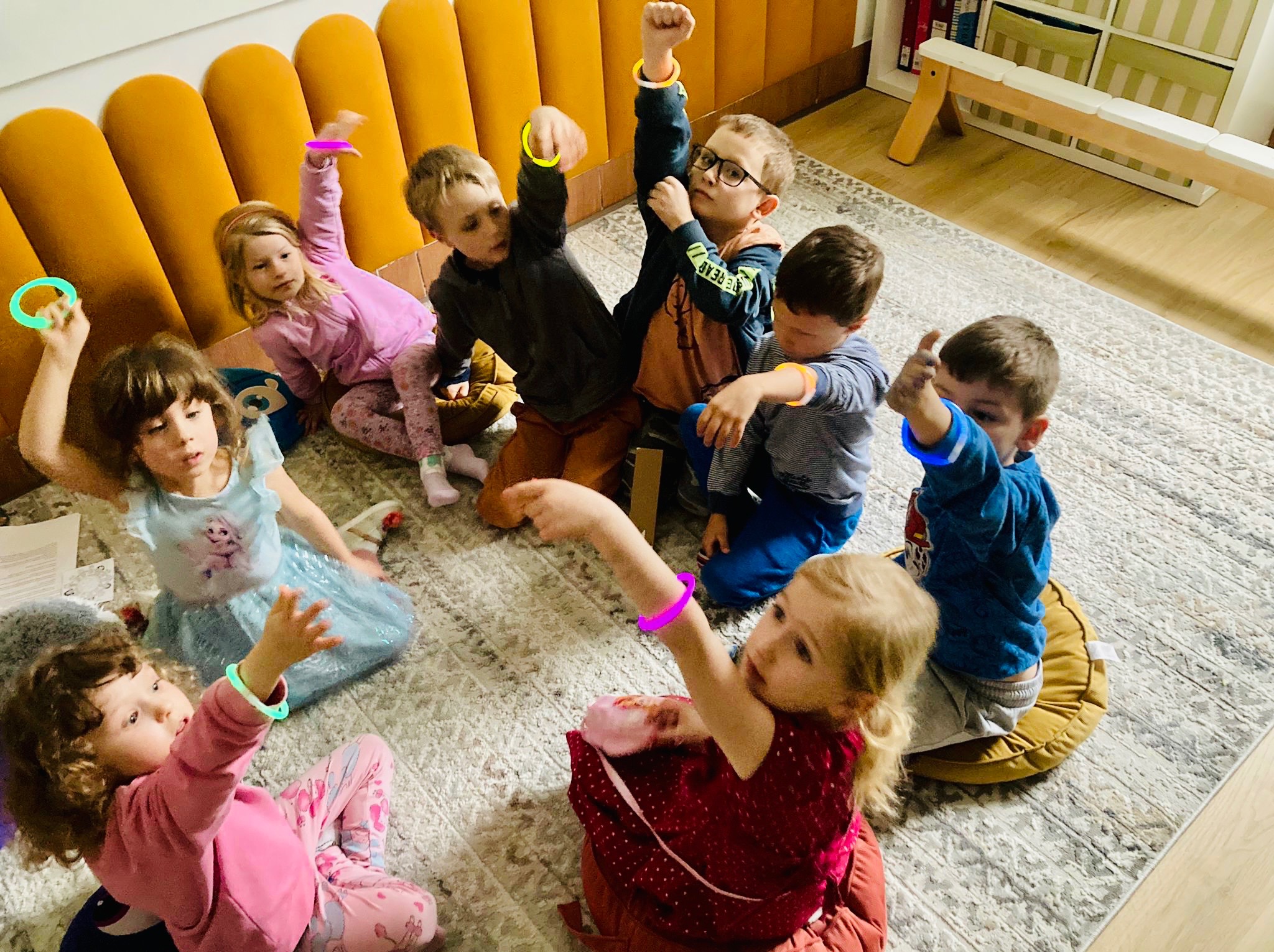 Grupa dzieci siedzi na dywanie w kole, z podniesionymi rękami i świecącymi bransoletkami, sugerując interaktywną zabawę lub zajęcia edukacyjne.