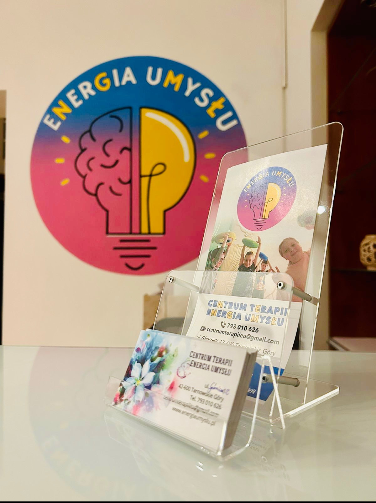Logo i wizytówki Centrum Terapii Energia Umysłu z motywem mózgu/żarówki, prezentowane w przezroczystym stojaku na blacie.