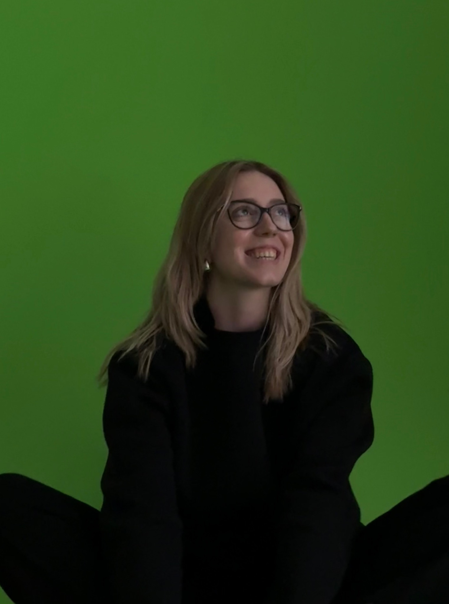 Uśmiechnięta blondynka w okularach, ubrana na czarno, siedzi na tle jaskrawozielonego ekranu, spoglądając w górę. Widoczny fragment sylwetki i twarz.