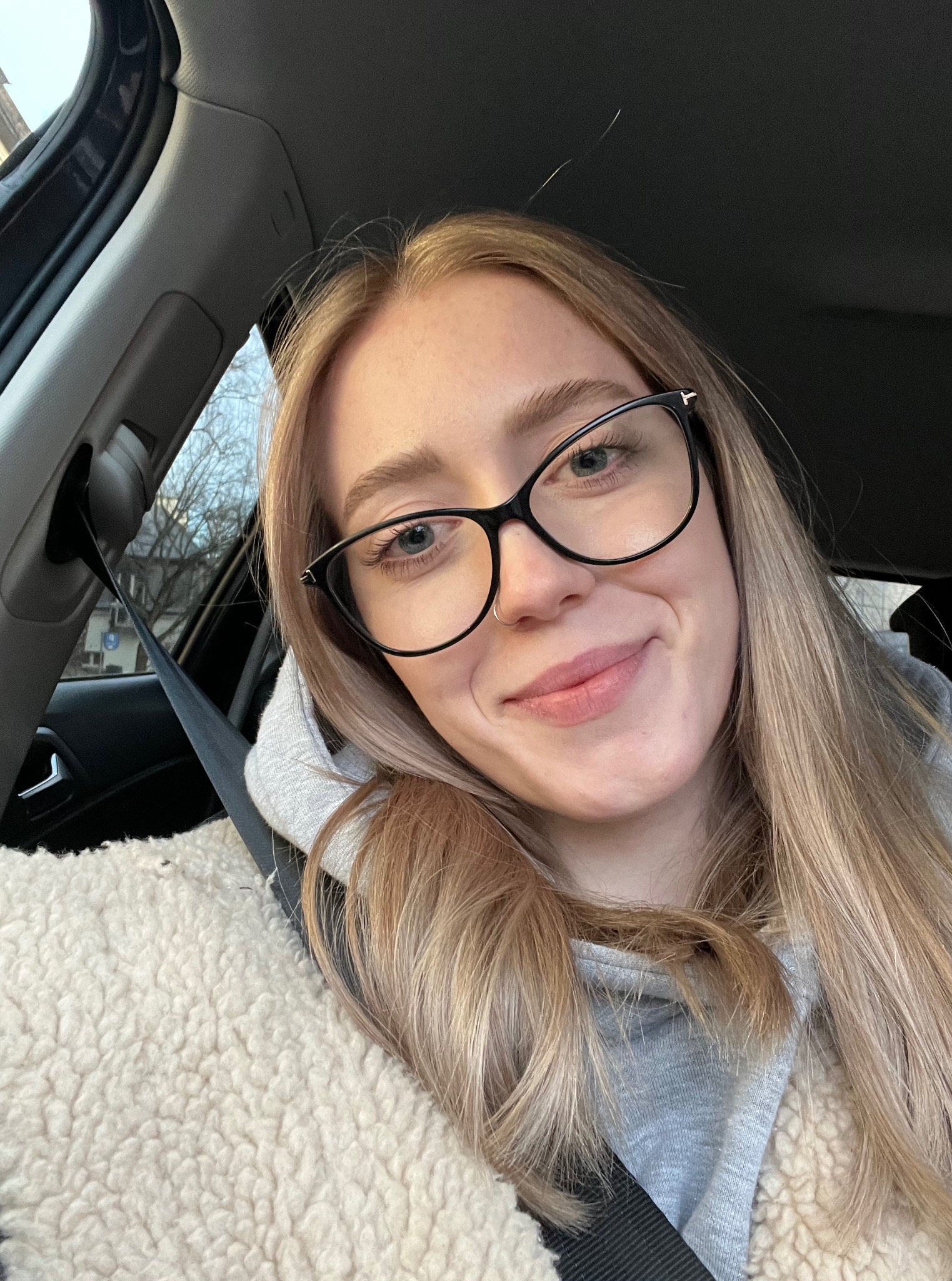 Uśmiechnięta blondynka w okularach w samochodzie, ubrana w szarą bluzę i białe futerko, zapięta pasem bezpieczeństwa. Widok z perspektywy pasażera.