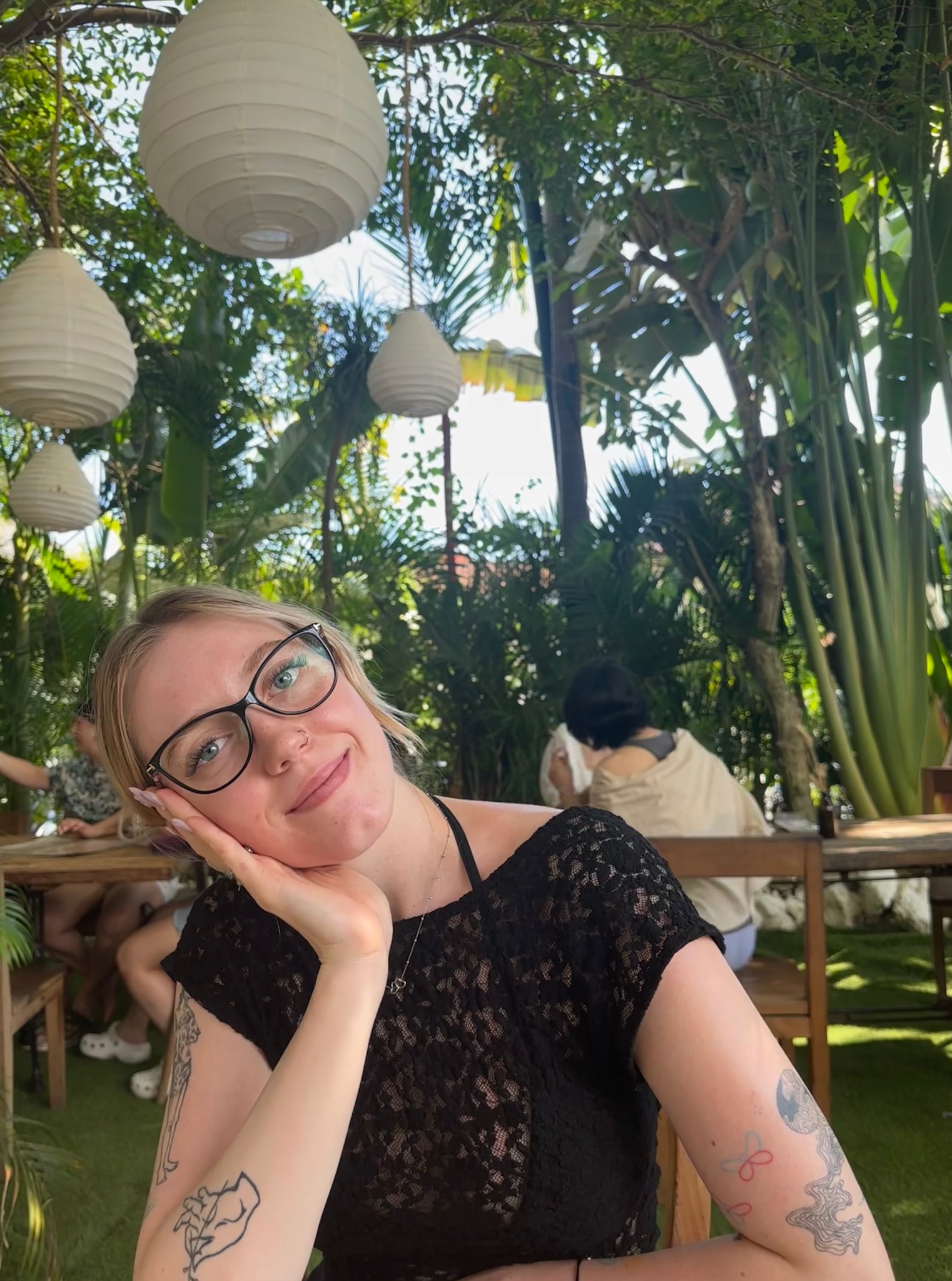 Portret młodej kobiety w okularach, opierającej głowę na dłoni, z widocznymi tatuażami na ramionach, w otoczeniu zieleni i lampionów w stylu boho.