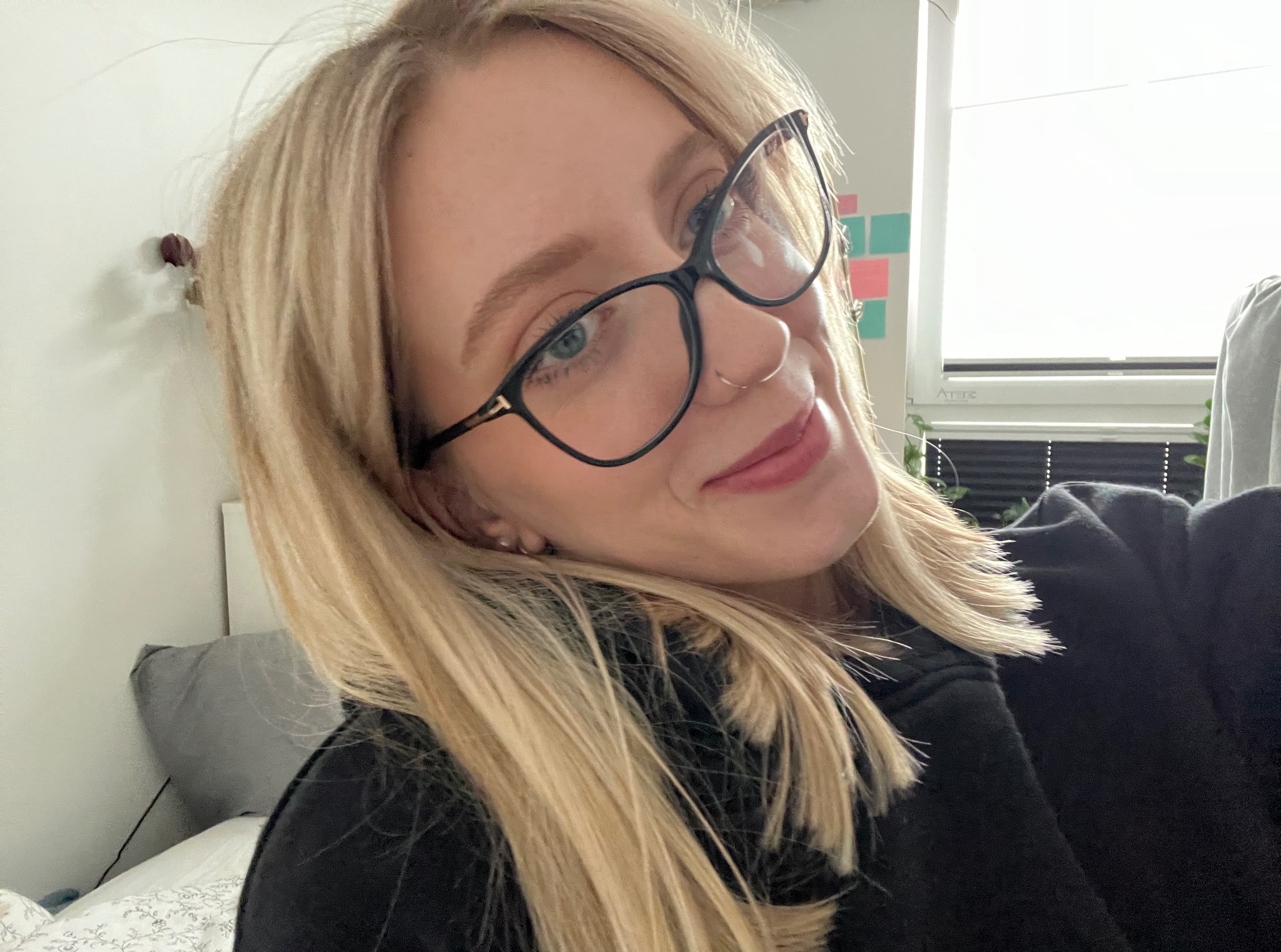 Portret młodej kobiety z blond włosami i okularami, uśmiechniętej, z kolczykiem w nosie, na tle pokoju z łóżkiem i oknem.