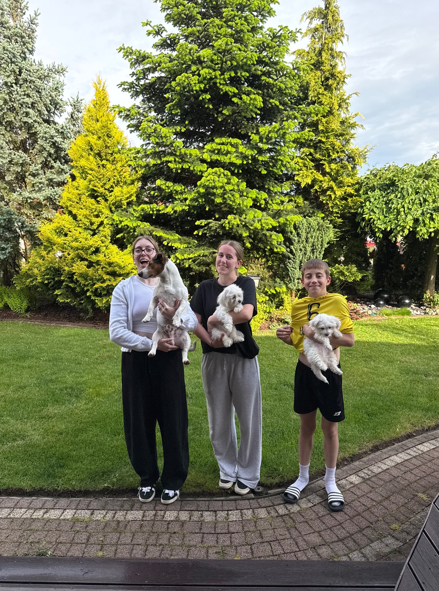 Trzy osoby trzymają małe psy na tle zielonego ogrodu z drzewami iglastymi i trawnikiem. Dziewczyna w okularach uśmiecha się, trzymając psa rasy Jack Russell Terrier.