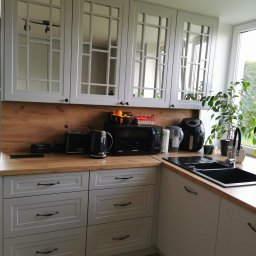 STOLARECZKA - Szara kuchnia na wymiar z drewnianym blatem i przeszklonymi szafkami wiszącymi. Widoczny zlew, bateria i sprzęty AGD. Uwagę zwraca doniczkowa roślina na blacie.