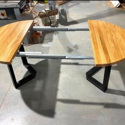 STOLARECZKA - Rozłożony, okrągły stół z drewnianym blatem i czarnymi, metalowymi nogami w kształcie trapezu. Widoczne mechanizmy rozsuwania blatu, maszyna stolarska w tle.