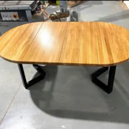 Rozkładany, owalny stół z jasnego drewna na czarnych, metalowych nogach w kształcie trapezu. Widoczne narzędzia stolarskie w tle.