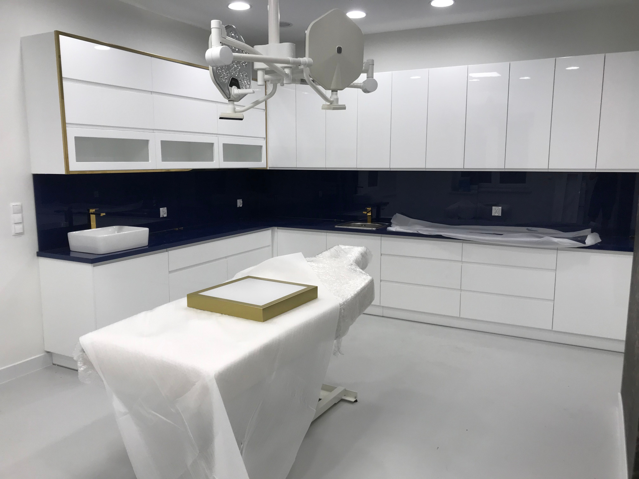 Białe, lakierowane szafki kuchenne z złotymi akcentami w sterylnym pomieszczeniu medycznym, z niebieskim blatem i sprzętem medycznym na pierwszym planie.