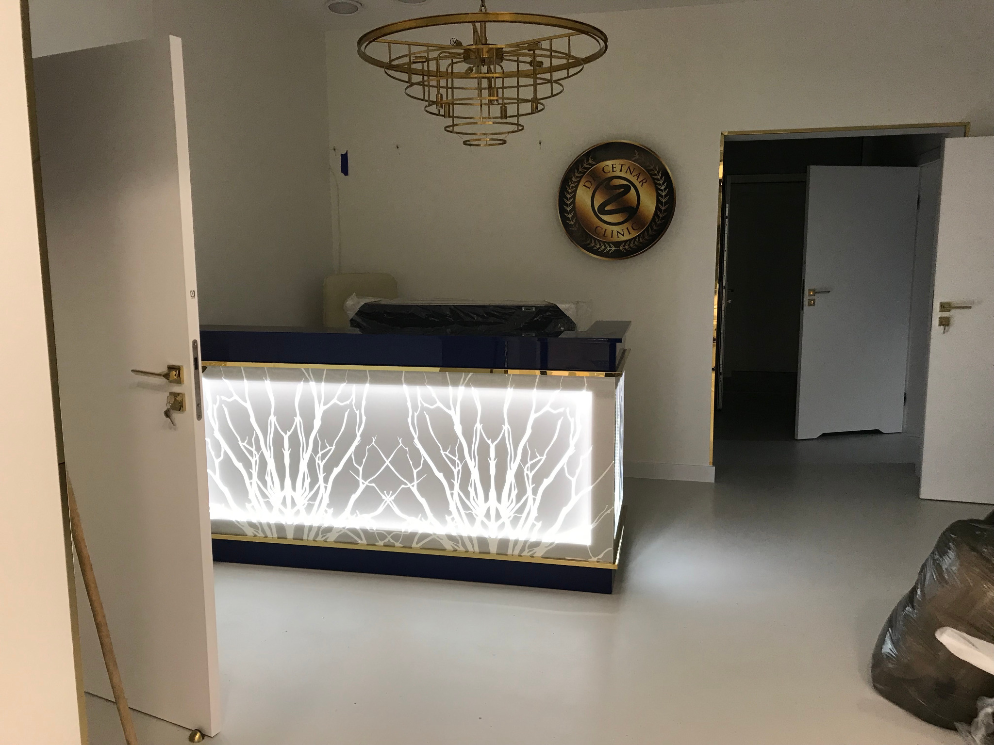 Nowoczesna lada recepcyjna z podświetlanym wzorem drzewa w nowym, minimalistycznym wnętrzu kliniki. Złoty żyrandol i logo na ścianie dodają elegancji.