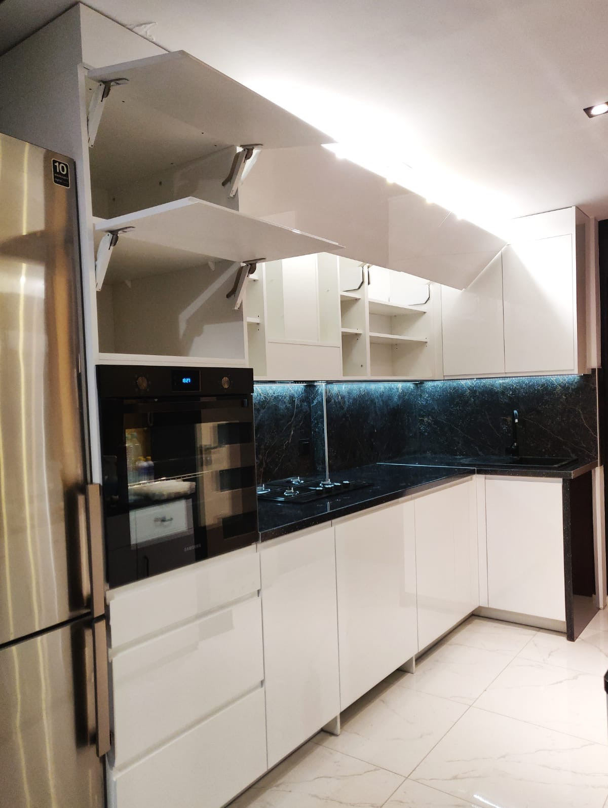 Biała kuchnia na wysoki połysk z czarnym blatem i otwartymi szafkami górnymi. Widoczny piekarnik i lodówka. Podświetlenie LED nad blatem.