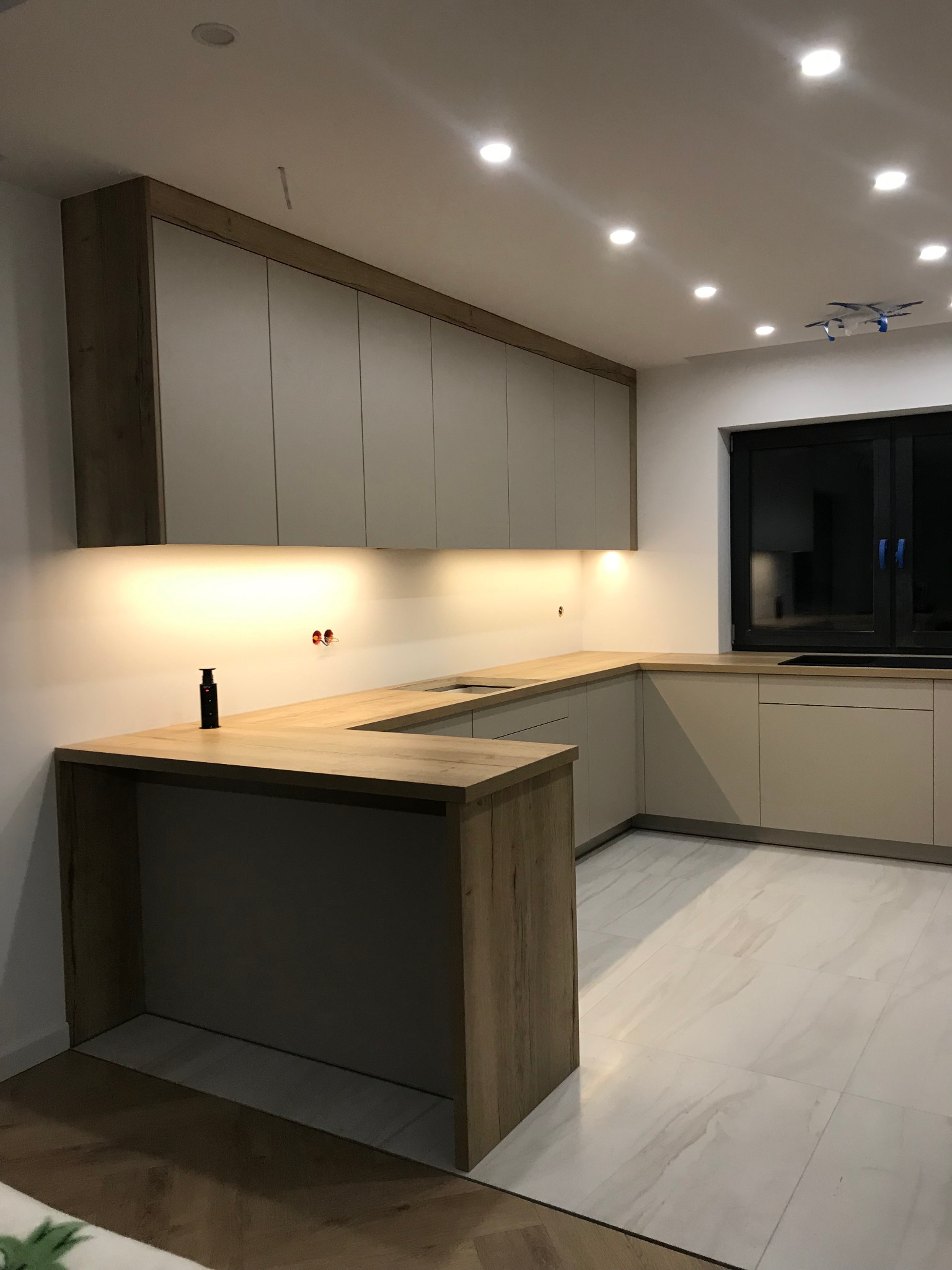 Jasna kuchnia w kształcie litery U z drewnianym blatem, oświetleniem podszafkowym i marmurową podłogą. Widoczne gniazdka elektryczne i wysuwany kontakt.