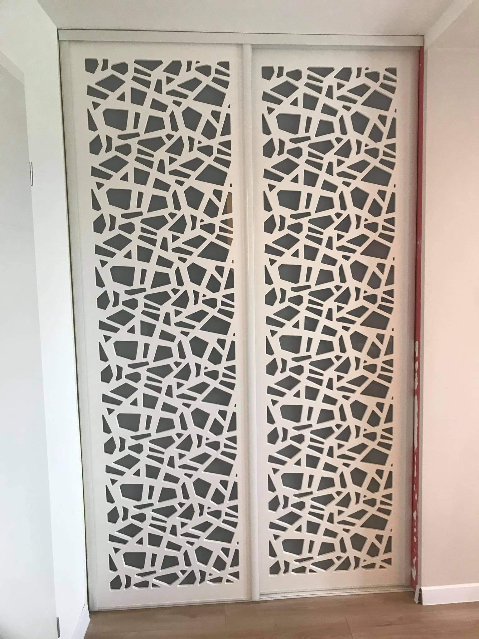 Białe, przesuwne drzwi szafy z abstrakcyjnym, geometrycznym wzorem na szarym tle, widoczne z bliska, z elementami wykończenia w kolorze czerwonym.