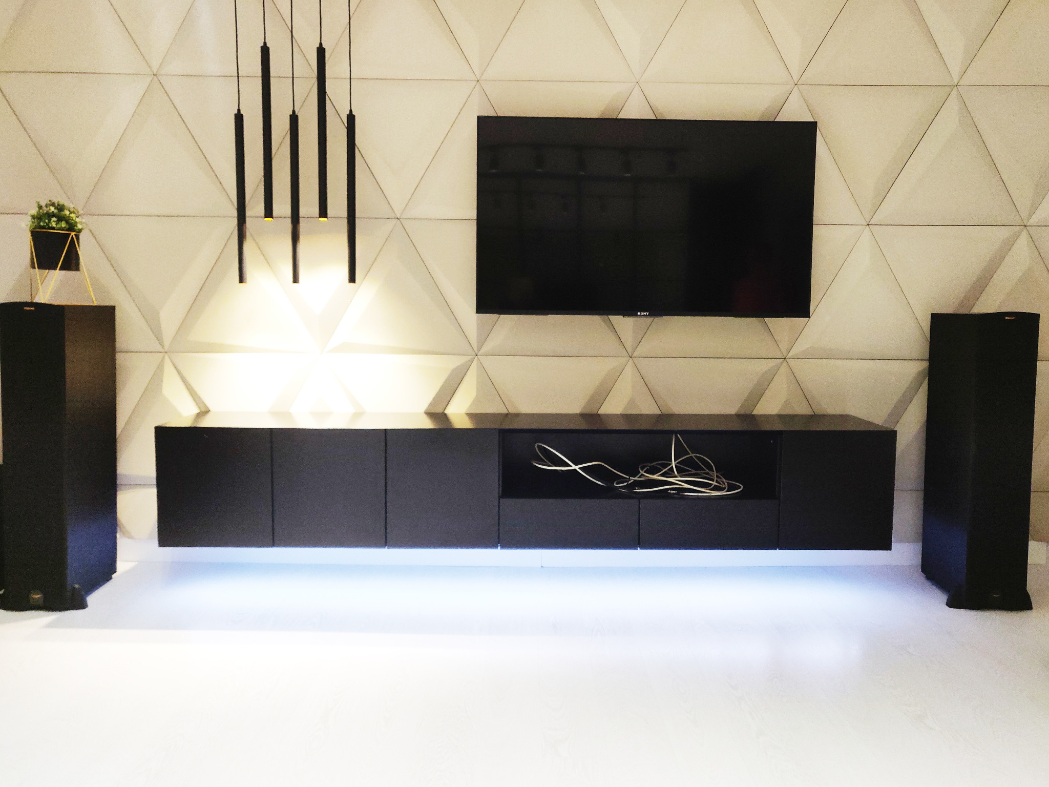 Minimalistyczna, czarna szafka RTV wisząca pod telewizorem, z podświetleniem LED. Na ścianie trójwymiarowa faktura i designerskie lampy wiszące.