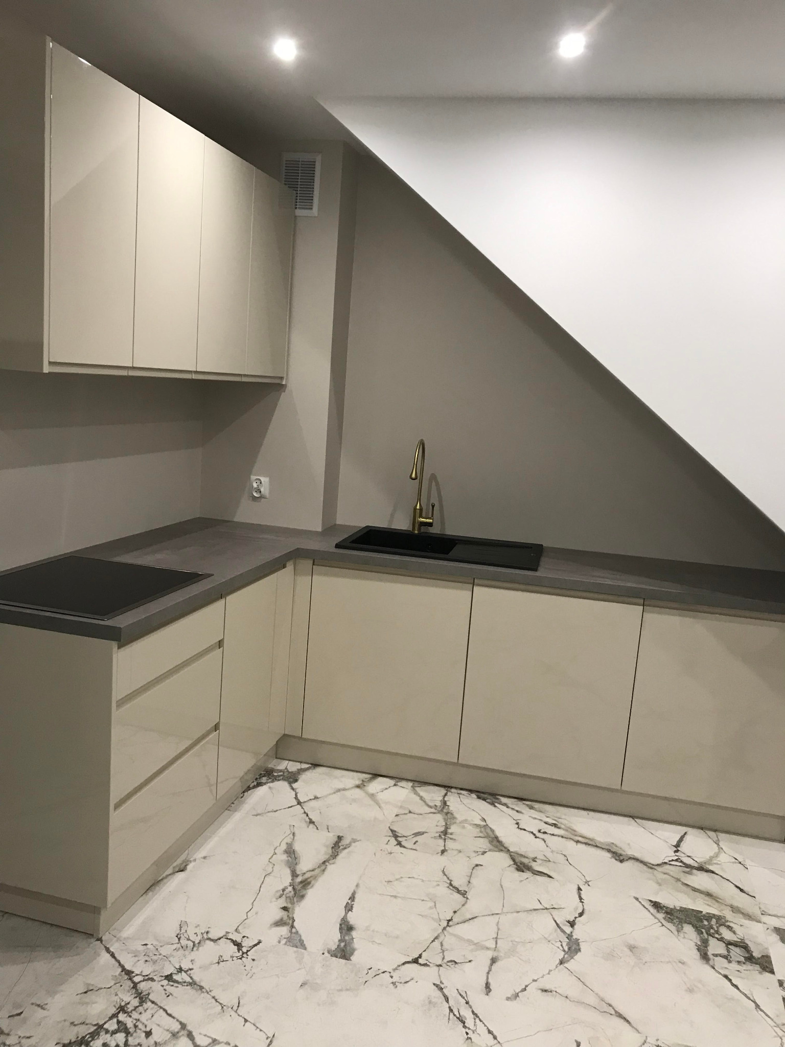 Jasna kuchnia z marmurową podłogą i złotą baterią. Zabudowa pod skosem dachu z płytą indukcyjną. Nowoczesny design z dbałością o szczegóły.