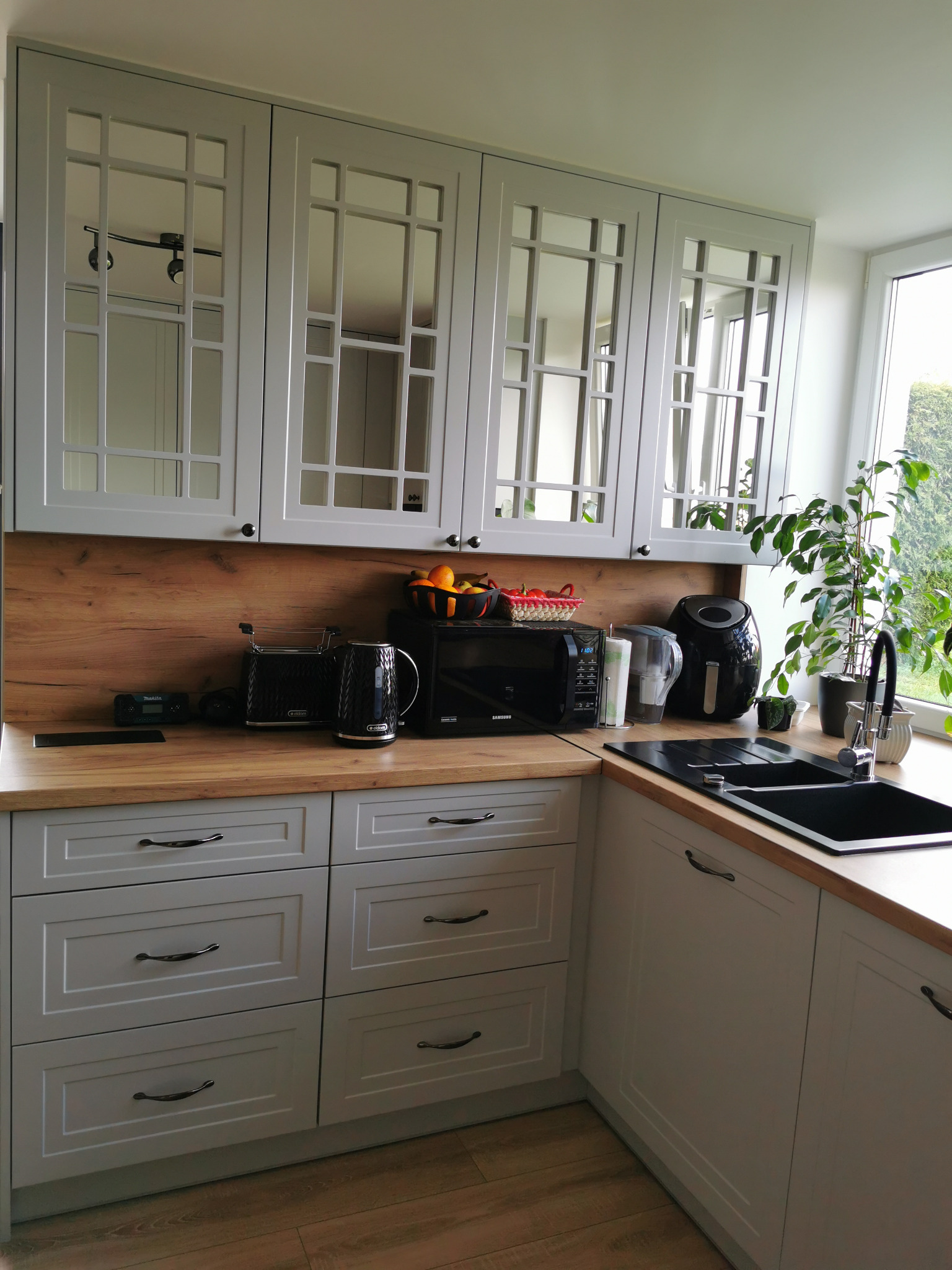 Szara kuchnia na wymiar z drewnianym blatem i przeszklonymi szafkami wiszącymi. Widoczny zlew, bateria i sprzęty AGD. Uwagę zwraca doniczkowa roślina na blacie.