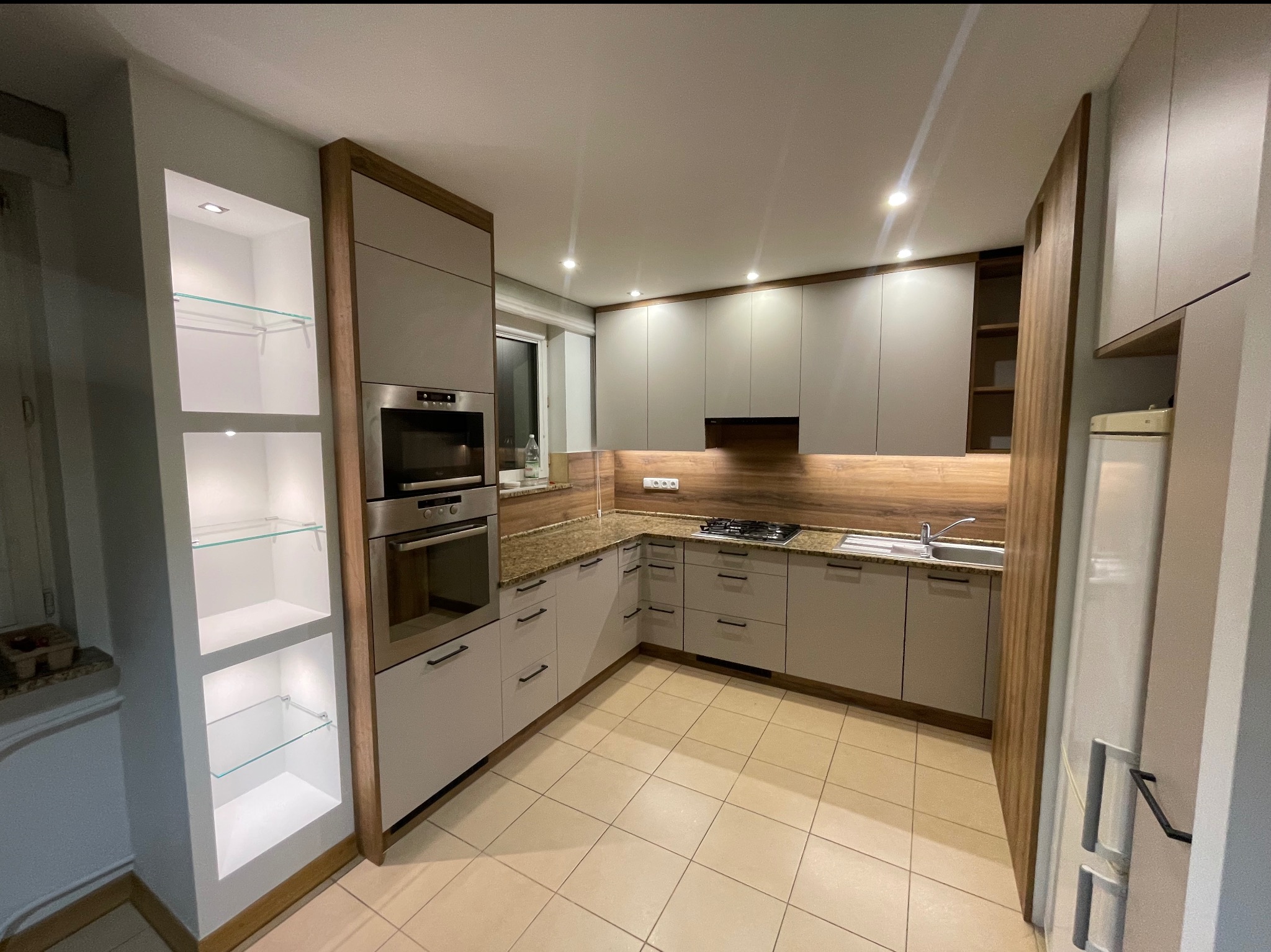 Jasna kuchnia z drewnianymi akcentami: zabudowa z piekarnikiem, płyta grzewcza, szklane półki z oświetleniem. Beżowe płytki podłogowe i drewnopodobny panel nad blatem.