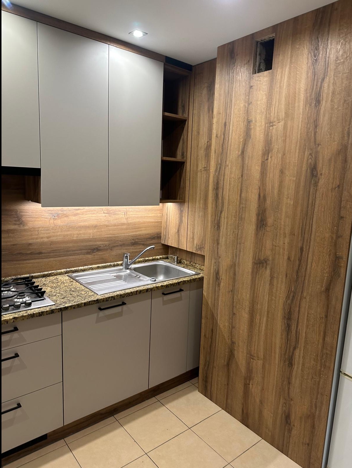 Jasna kuchnia na wymiar z drewnianymi akcentami: szafki górne w odcieniach szarości, blat z efektem kamienia i fronty imitujące drewno. Widoczna płyta grzewcza i zlew.