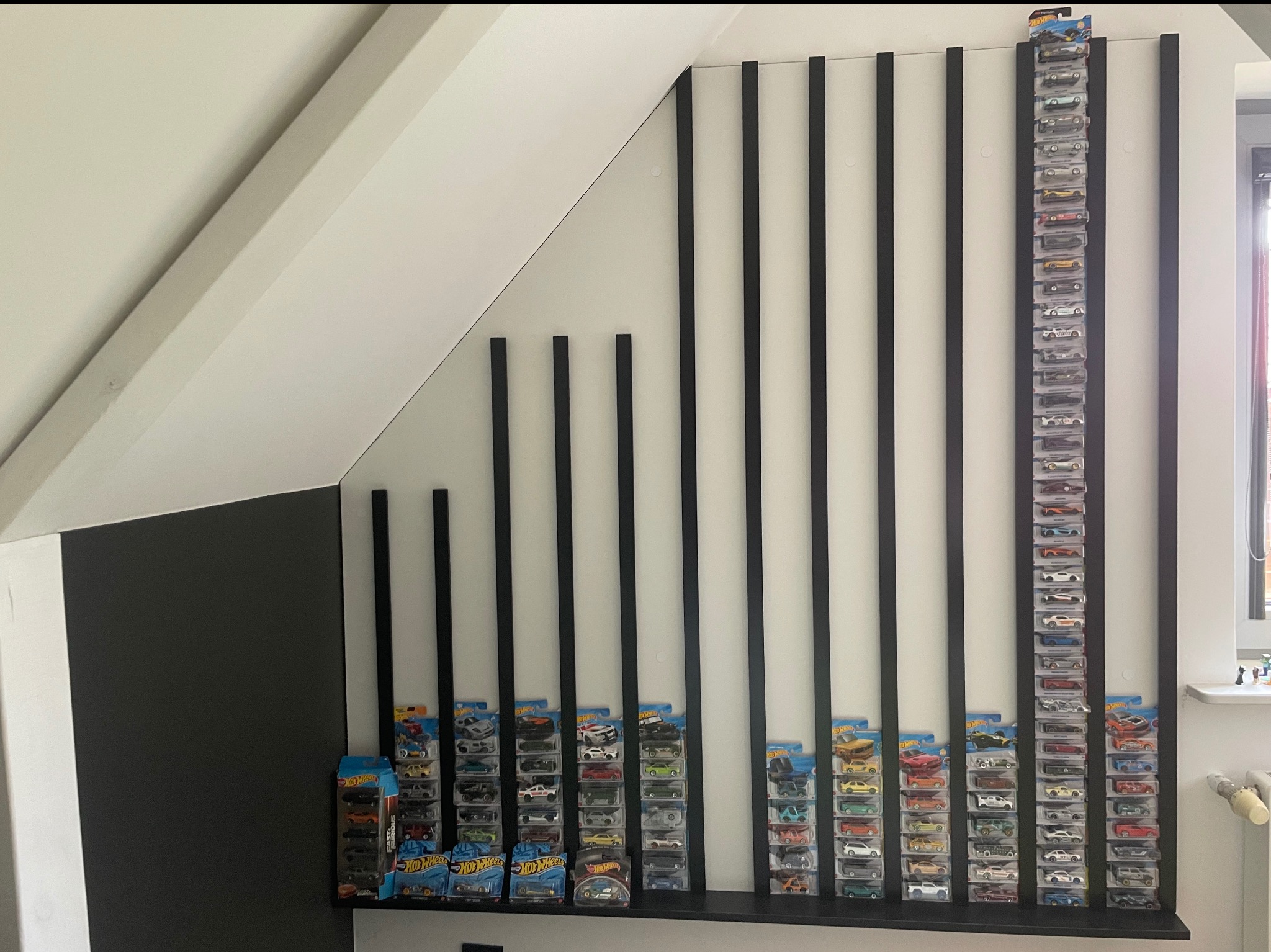 Nietypowa półka na kolekcję autek Hot Wheels, czarne pionowe listwy na białej ścianie pod skosem, zabawki w oryginalnych opakowaniach.