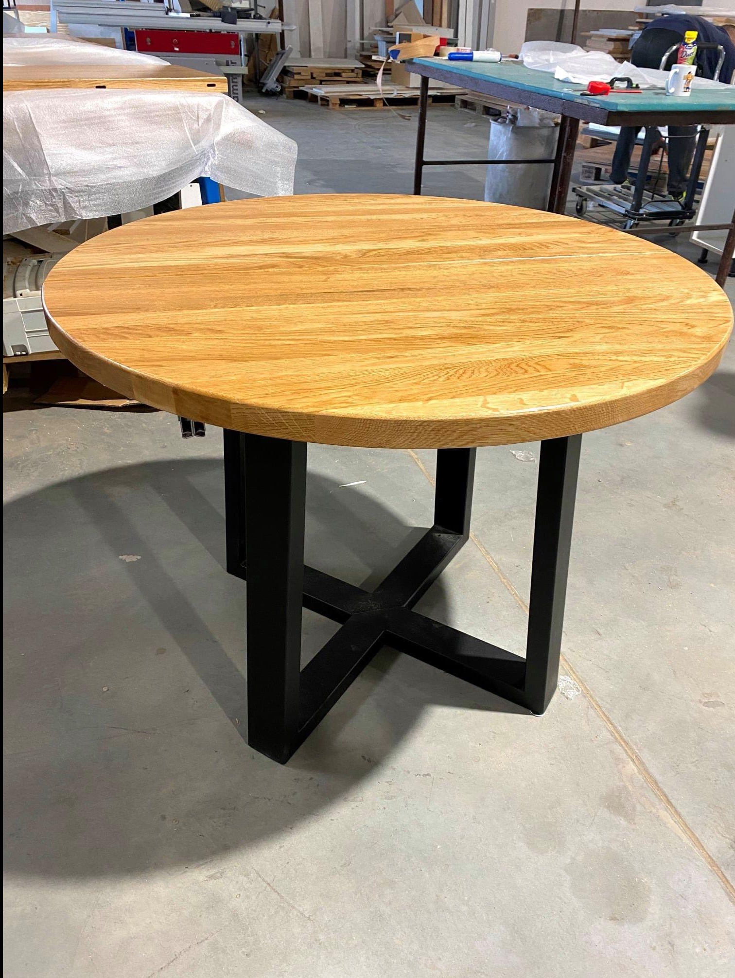 Okrągły stół z blatem z jasnego drewna i czarną, metalową podstawą w kształcie litery X, widoczny w warsztacie stolarskim.