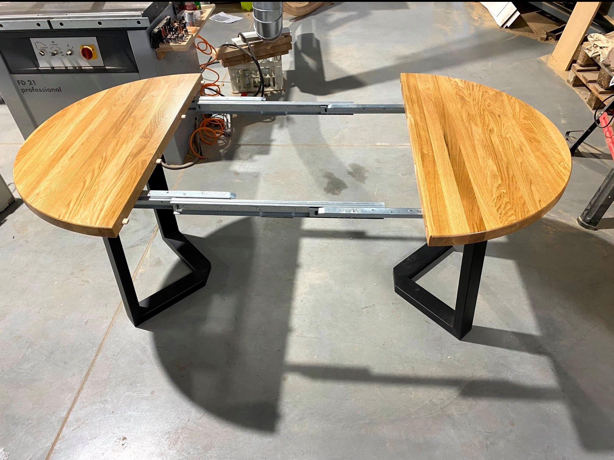 Rozłożony, okrągły stół z drewnianym blatem i czarnymi, metalowymi nogami w kształcie trapezu. Widoczne mechanizmy rozsuwania blatu, maszyna stolarska w tle.