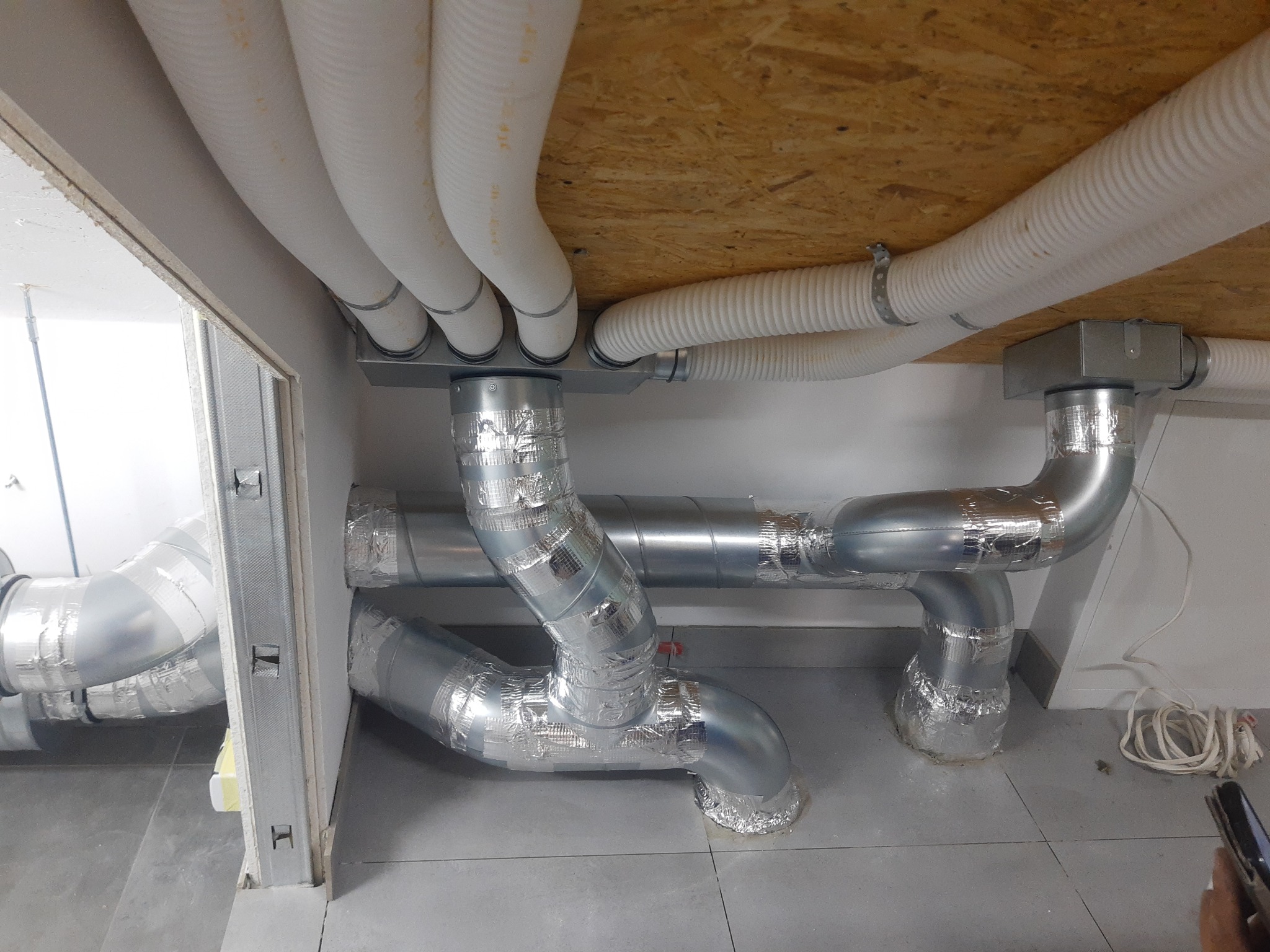 Instalacja wentylacyjna z białymi i srebrnymi rurami pod sufitem z płyty OSB, widoczne łączenia zabezpieczone taśmą aluminiową.