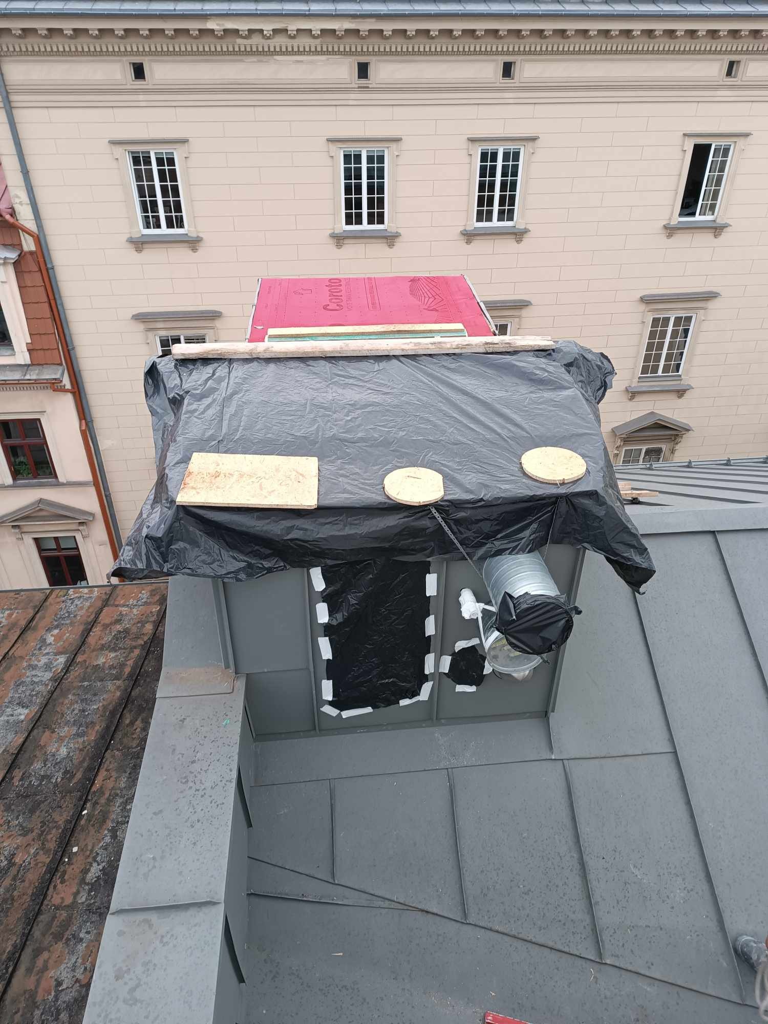 Prace na dachu budynku z wentylacją, zabezpieczone folią i deskami. Widoczne elementy instalacji wentylacyjnej i materiały budowlane. W tle budynek z oknami.