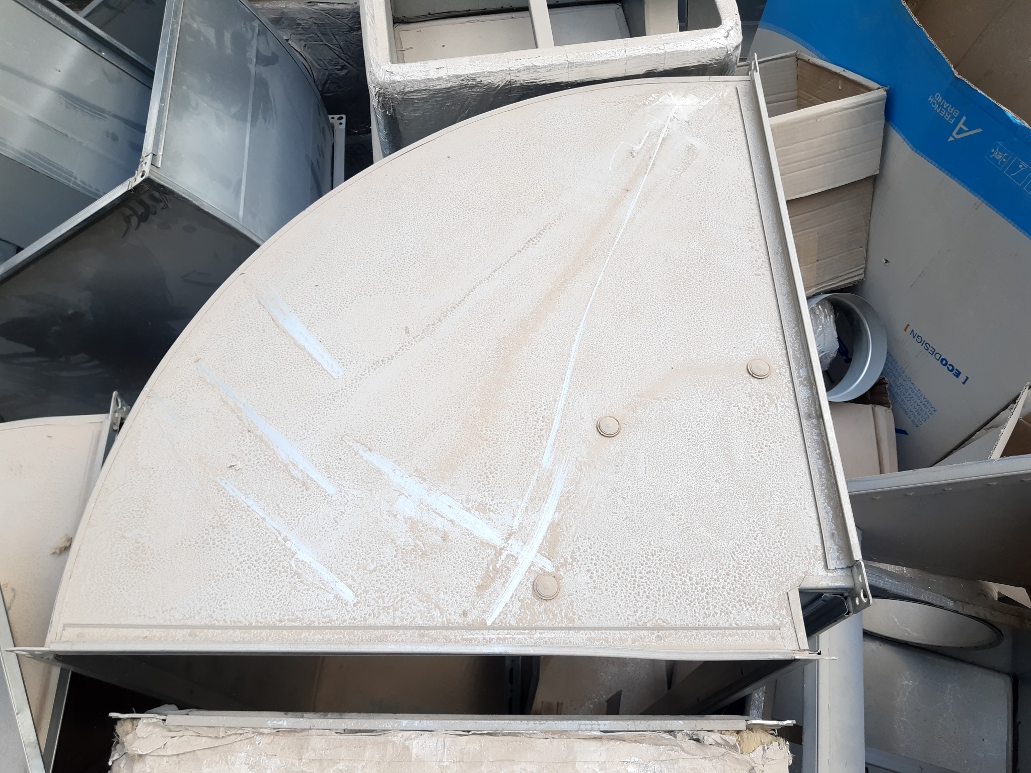 Kątowy element wentylacji, pokryty warstwą kurzu, otoczony innymi częściami instalacji i kartonami. Widoczne ślady po czyszczeniu na powierzchni.