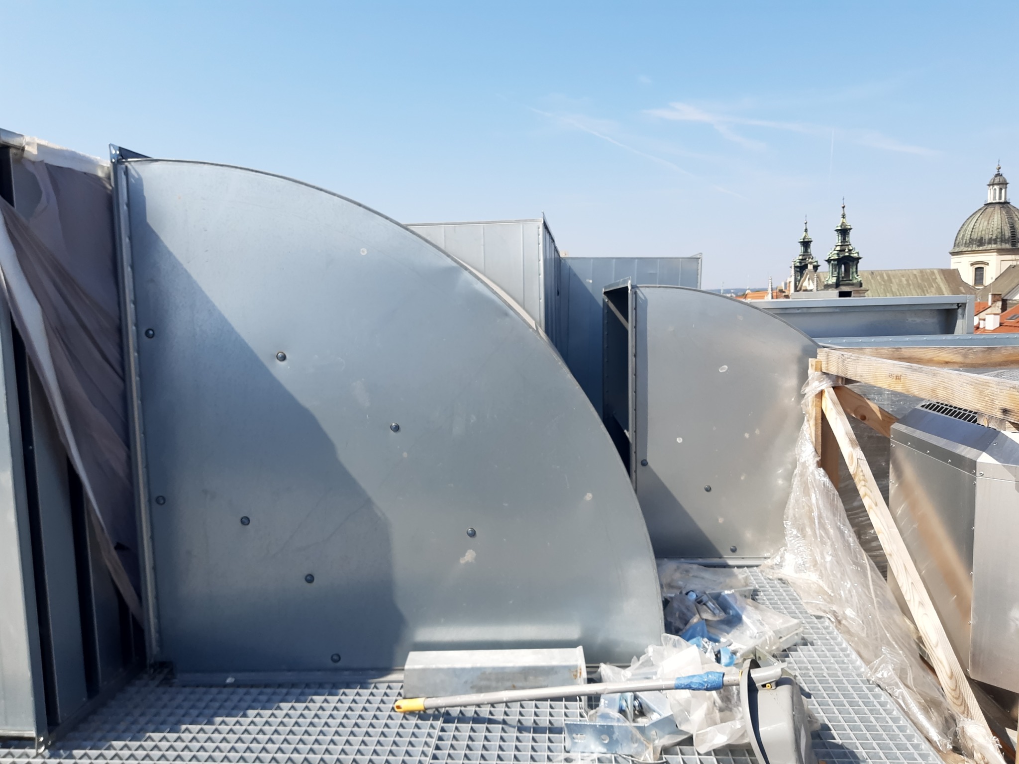 Instalacja kanałów wentylacyjnych na dachu z widokiem na budynki sakralne w tle. Elementy w trakcie montażu, widoczne narzędzia i folia ochronna.