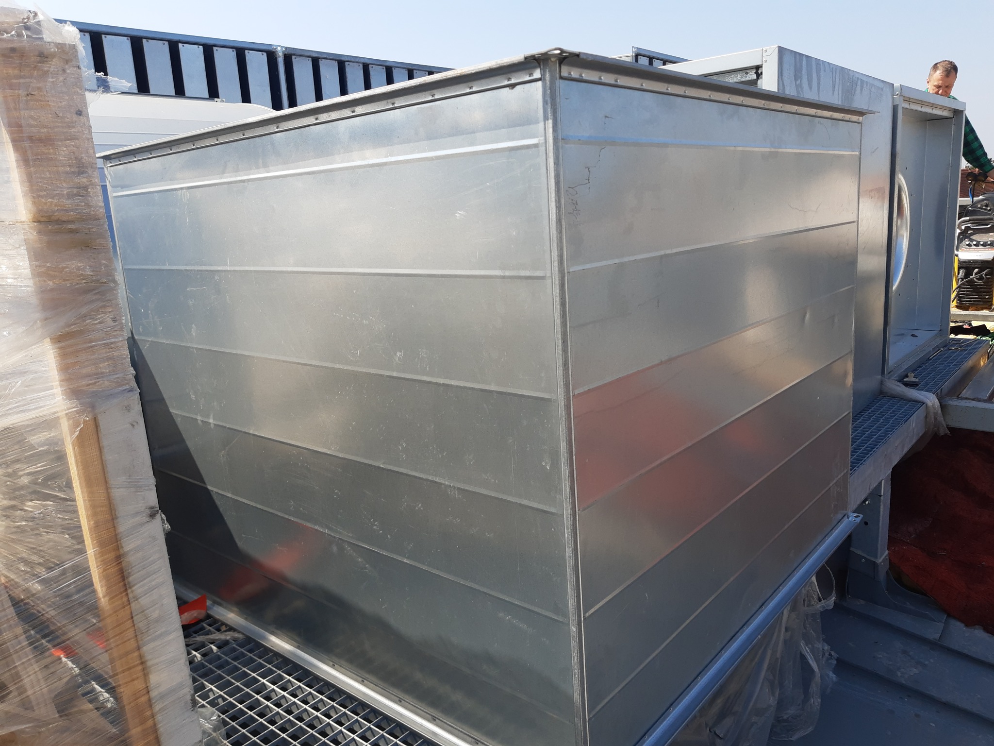 Montaż przemysłowego wentylatora dachowego z blachy ocynkowanej. Widoczny fragment dachu i osoba nadzorująca instalację. Jasne oświetlenie dzienne.