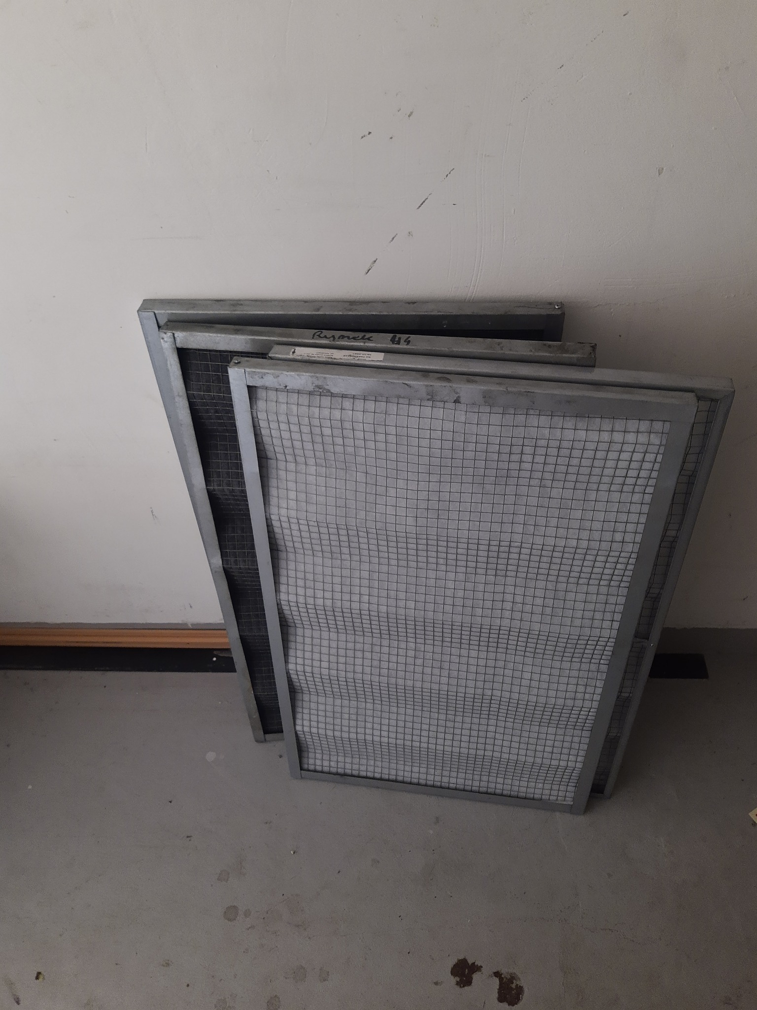 Stos filtrów wentylacyjnych z metalową ramą, ustawionych pod kątem przy białej ścianie. Widoczne ślady użytkowania i napisy na filtrach.