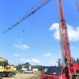 Czerwony dźwig samojezdny Spierings SK599-AT5 na placu budowy, w tle żółta koparka CAT M316F i samolot na błękitnym niebie.