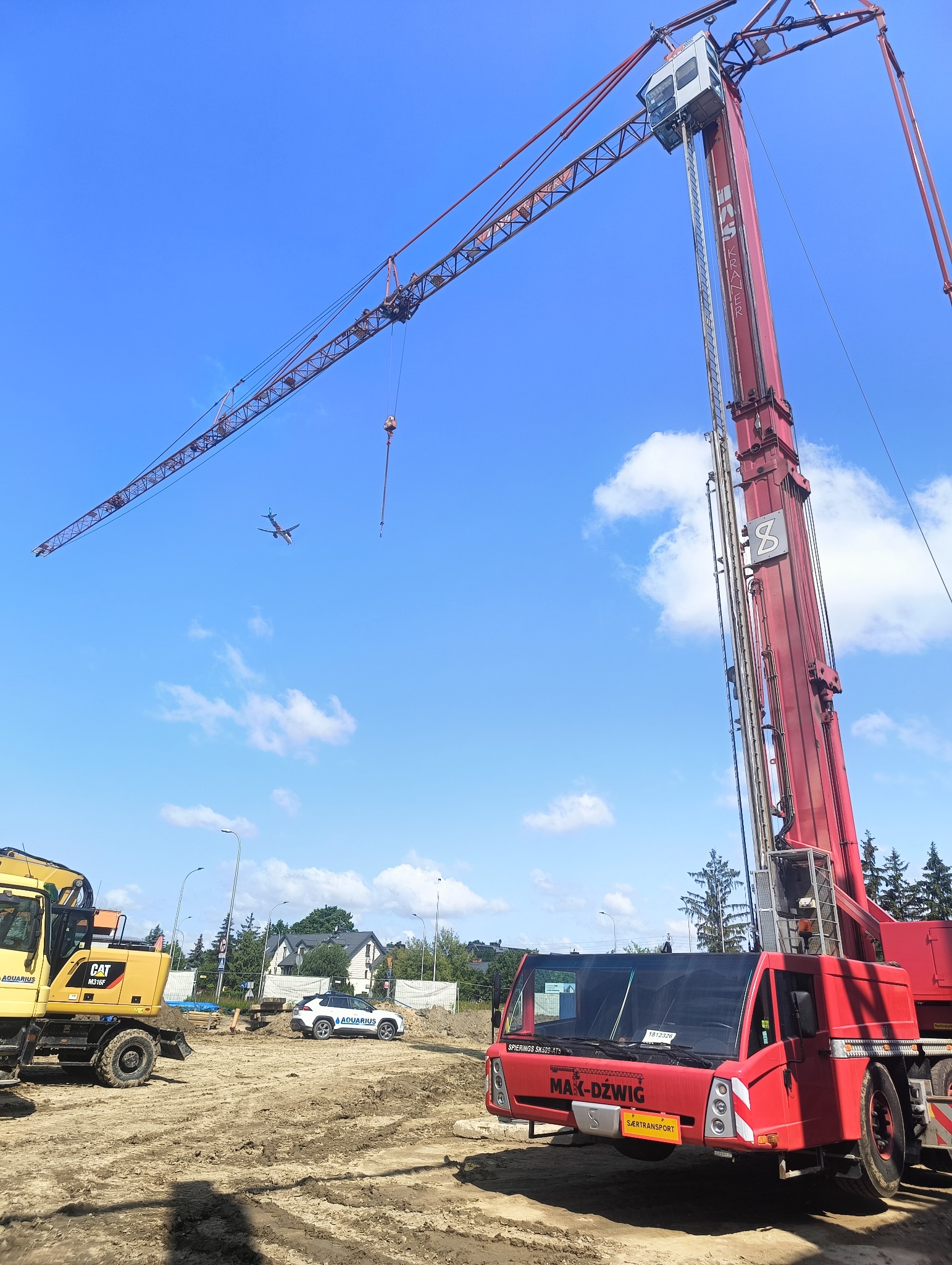 Czerwony dźwig samojezdny Spierings SK599-AT5 na placu budowy, w tle żółta koparka CAT M316F i samolot na błękitnym niebie.
