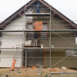 ON BUD Michał Zejer - Dom w trakcie ocieplania: jedna ściana z izolacją, druga z nałożonym tynkiem. Ekipa na rusztowaniu wykonuje prace wykończeniowe elewacji.